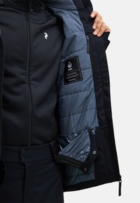 Schwarze Außenjacke mit einer blau gesteppte Innenfutter, die ein Logo, Reißverschlüsse und Taschen aufweist. Das Material wirkt wasserabweisend und robust.