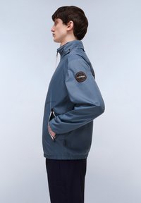 Blauwe waterdichte jas met een hoge kraag, ritssluitingen in de zakken en een logo patch op de linkermouw. Glad textuur en elastische zoom.