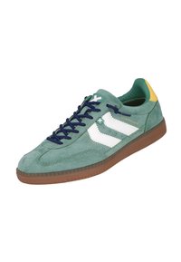 Zapatilla de ante verde con rayas blancas, cordones azul marino y un acento amarillo en el talón. Presenta una suela de goma marrón texturizada.