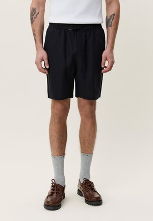 Man draagt zwarte short met trekkoord, grijze crew-sokken, bruine leren schoenen en een wit mouwloos shirt, staand met ontspannen armen.
