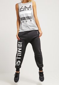Canotta grigia senza maniche con stampa "CITY WARRIOR", abbinata a pantaloni jogger neri con testo bianco "LES MILLS". Scarpe da atletica nere.