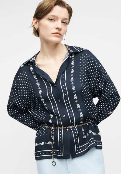 Νεαρό άτομο με κοντά μαλλιά φοράει ένα πουκάμισο σε σκούρο μπλε με σχέδια και κουμπιά, ζωνασμένο στη μέση με μια χρυσή αλυσίδα-ζώνη.