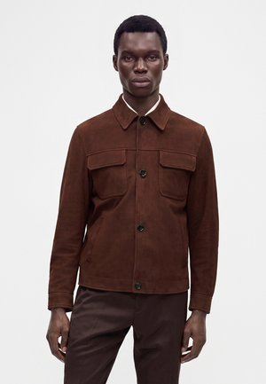 Mann trägt eine dunkelbraune Wildlederjacke mit Knöpfen und zwei Brusttaschen sowie passende braune Hose vor einem schlichten weißen Hintergrund.