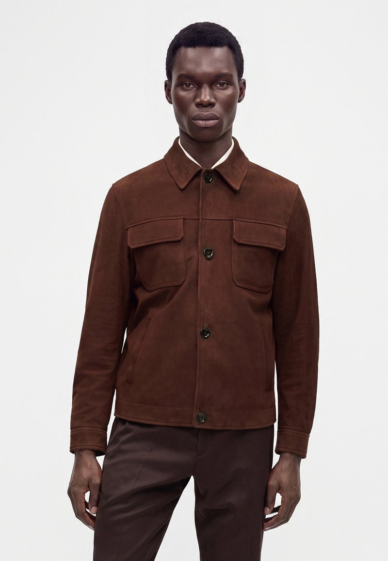 Homme portant une veste en daim marron foncé boutonnée avec deux poches poitrine et un pantalon marron assorti sur un fond blanc uni.