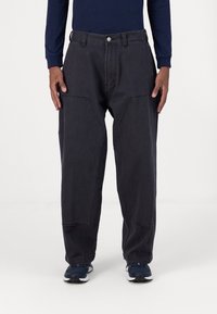 Black, loose-fit cargo pants with large side pockets, visible stitching, and a button closure. Worn with blue athletic shoes.