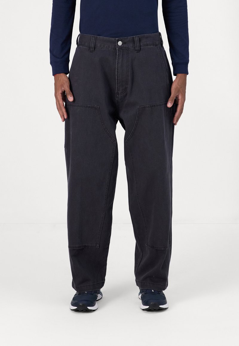 Black, loose-fit cargo pants with large side pockets, visible stitching, and a button closure. Worn with blue athletic shoes.