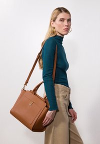 Bolso de mano rectangular de cuero marrón con asas dobles y correa ajustable para el hombro. Textura suave y un bolsillo frontal con un pequeño emblema.