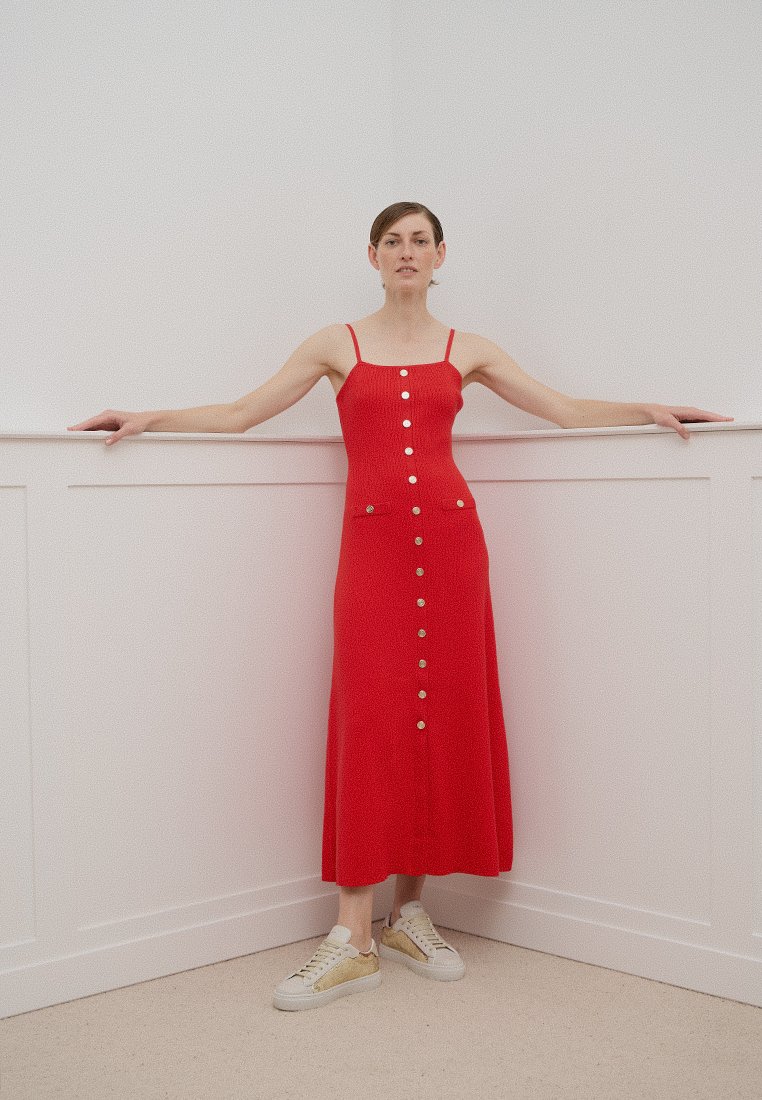 Vestido midi rojo sin mangas con un corsé ajustado, detalles de botones dorados en la parte delantera y un dobladillo acampanado, combinado con zapatillas blancas y doradas.