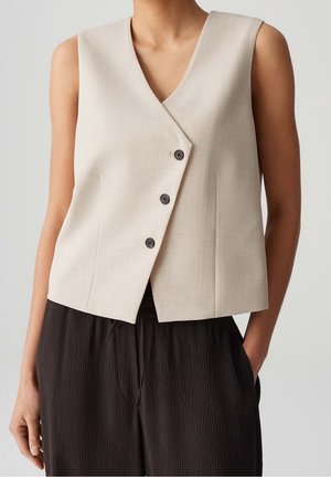 Gilet beige sans manches avec trois boutons porté sur un pantalon noir texturé sur une personne avec une main dans la poche.
