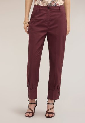 Femme portant un pantalon bordeaux taille haute et fuselé avec des poignets boutonnés et des sandales à talons hauts noires à brides, debout devant un fond uni.