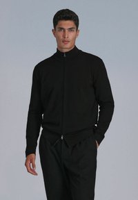 Veste noire zippée sur le devant en tissu tricot lisse, avec un col haut, des manches longues et des coutures marquées au niveau de la taille.