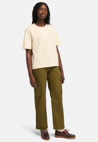 Krémové bavlněné tričko s krátkým rukávem a čtvercovým logem. Olivové cargo kalhoty s bočními kapsami. Hnědé boty s texturovanou podrážkou.
