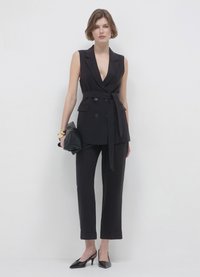 Gilet su misura nero con scollo a V profondo, davanti doppiopetto, vita con cintura; abbinato a pantaloni neri a vita alta e a sigaretta e tacchi a punta.