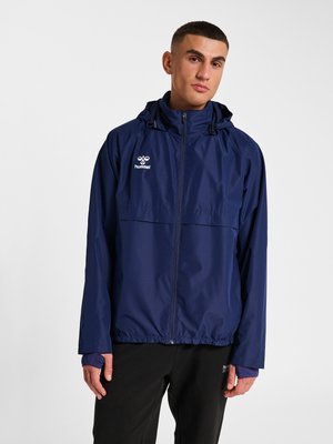 Joven con chaqueta con capucha azul marino y cremallera y pantalones negros, de pie frente a un fondo claro y liso.