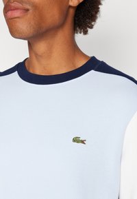Felpa azzurra chiara con bordi blu navy e un logo di coccodrillo verde ricamato sul petto. Il tessuto sembra liscio con una vestibilità casual.