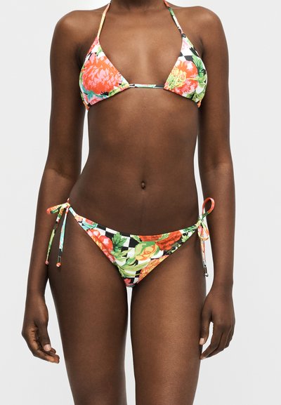 Bikini triangle à imprimé floral avec des fleurs orange, vertes et jaunes sur un fond rayé noir et blanc, porté par une personne debout.
