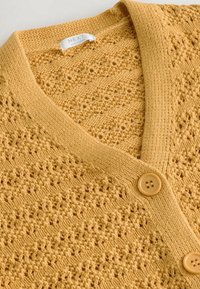 Cardigan jaune tricoté avec col en V, fermeture par boutons et motif ajouré texturé, étiqueté "NEXT Made in China."
