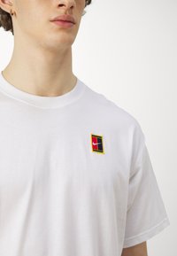 T-shirt bianca in cotone con una piccola patch logo con design geometrico rosso, nero e giallo sulla parte sinistra del petto.