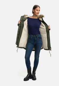 Olivengrønn parkas med pelsbesatt hette, beige plysjfor, kombinert med en marineblå topp og blå skinny jeans, fullført med svarte ankelstøvletter.