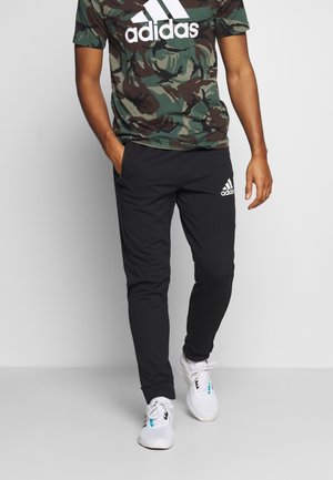 Mężczyzna w czarnych spodniach Adidas z białym logo, koszulka Adidas z nadrukiem moro i białe buty sportowe, ręce częściowo w kieszeniach, stoi przy szarej ścianie.