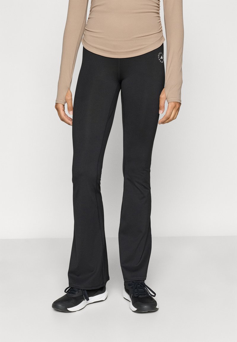 Μαύρα κολάν με φαρδιά καμπάνα, κατασκευασμένα από ελαστικό υλικό, με λεία υφή και μικρό λογότυπο κοντά στη μέση. Τα παπούτσια είναι μαύρα αθλητικά με λευκές σόλες.