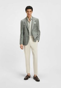Veste à carreaux gris clair avec revers en pointe, chemise beige et pantalon crème. Des mocassins marron complètent l'ensemble, mettant en valeur la coupe ajustée et les textures.