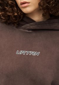 Καφέ φούτερ με κουκούλα από μαλακό ύφασμα, με τσέπη καγκουρό μπροστά και μεγάλο λευκό λογότυπο "URTRN" στο στήθος.