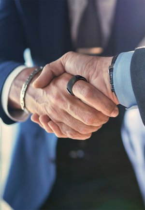 Deux mains se serrent, l'une avec une bague noire lisse et l'autre avec un bracelet en argent. Les deux portent une tenue formelle avec une chemise bleue et un costume.