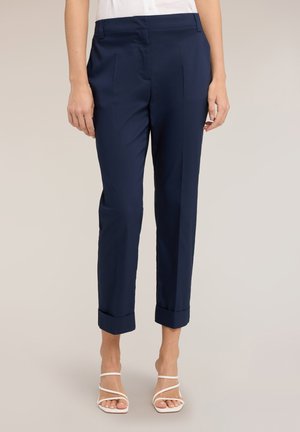 Kvinde iført skræddersyede navyblå ankellange bukser med manchetter, kombineret med hvide sandaler med hæl og stropper på en ensfarvet baggrund.