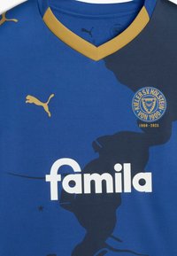 Modrá športová dres s výstrihom do V, zlatými akcentmi, veľkým bielym logom "famila" a kruhovým odznakom Kieler SV Holstein na ľavej strane.