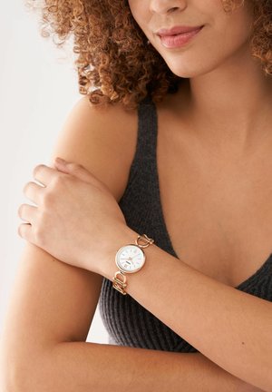 Vrouw met krullend haar, gekleed in een donker mouwloos topje en een gouden horloge met een witte wijzerplaat aan haar linkerpols, die haar rechterarm vasthoudt.