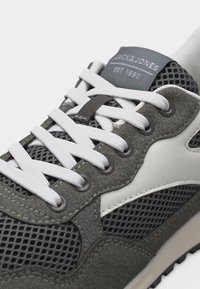 Nahaufnahme eines grauen und weißen Mesh-Sportschuhs mit weißen Schnürsenkeln und einem "JACK & JONES EST. 1990" Etikett auf der Zunge.