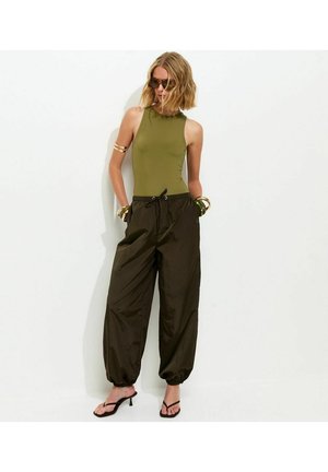 Femme portant un haut sans manches vert olive, un pantalon ample foncé avec un cordon, des sandales à talons noires, des bracelets dorés et des lunettes de soleil sur fond blanc.