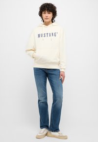 Crème hoodie met "MUSTANG" logo, capuchon met trekkoord, voorzak; gecombineerd met blauwe jeans en lichtgekleurde sneakers.