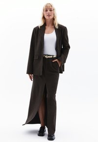 Blazer marrón ajustado con un solo botón, combinado con una falda larga con una abertura frontal, top blanco sin mangas y un cinturón metálico. Zapatos negros.