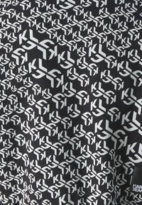 Černá látka s opakujícím se bílým vzorem písmen 'K' a 'L'. Design je geometrický a hustý, s hladkým povrchem.