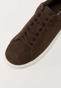 Brown suede sneaker with a rounded toe, flat laces, and a white rubber sole. Upper features a smooth texture and minimal branding.