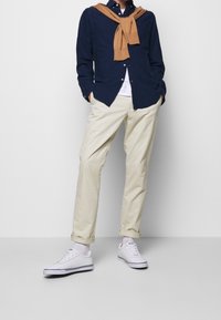 Marinblå skjorta, beige chinos, vita sneakers med svarta accenter och en beige stickad tröja hängande runt nacken. Avslappnad outfit.
