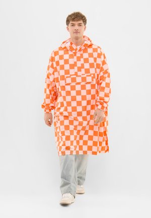 Mladý muž v oranžovo-bílé kostkované volné kapucové bundě, světle modrých džínách a bílých teniskách kráčí vpřed na bílém pozadí.