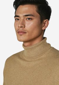 Beiger Rollkragenpullover aus weichem Strickstoff, mit hohem Kragen und gerippten Kanten, präsentiert an einem Modell vor weißem Hintergrund.
