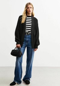 Zwarte gebreide cardigan over een zwart-wit gestreept shirt, gecombineerd met blauwe flared jeans en zwarte sneakers, terwijl een kleine zwarte tas wordt vastgehouden.