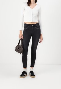 Schwarze hochtaillierte Jeans mit Strassakzenten, kombiniert mit einem weißen, gerippten Langarm-Crop-Top und schwarzen Sneakers. Kleine braune Handtasche.