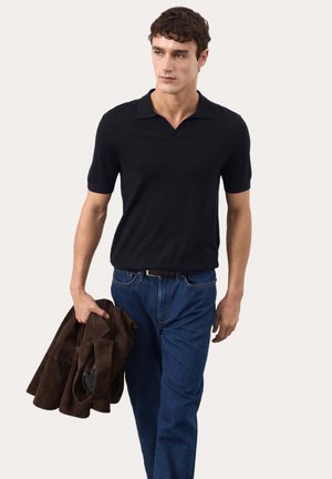 Giovane uomo che cammina indossando una polo nera a maniche corte e jeans blu, con in mano sinistra una giacca marrone piegata.