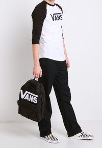 Schwarz-weißer Vans-Logo-Rucksack, schwarze Hosen und weiße Schuhe mit kariertem Muster. Der Mann trägt ein schwarz-weißes Vans-Raglan-Shirt.