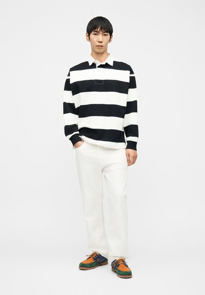 Άνδρας που στέκεται με τα χέρια στις τσέπες, φοράει μακρυμάνικο πουκάμισο με μαύρες και λευκές φαρδιές ρίγες, λευκό παντελόνι και πολύχρωμα casual παπούτσια.