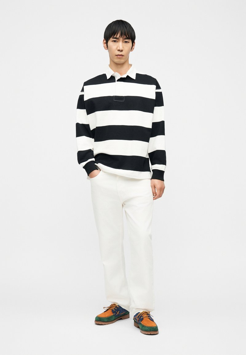 Homme debout, les mains dans les poches, portant une chemise à manches longues à larges rayures noires et blanches, un pantalon blanc et des chaussures décontractées multicolores.