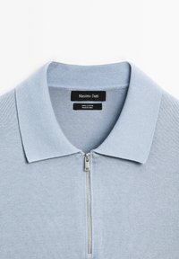 Polo en coton bleu clair avec une demi-fermeture éclair argentée et des étiquettes noires indiquant la marque et les détails du tissu.