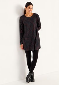 Kvinna lutad mot vit vägg, klädd i en svart tunika med röda vågiga linjer, svarta leggings och svarta ankelstövlar, leende och tittande åt höger.