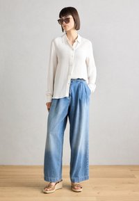 Chemise blanche à manches longues côtelées avec des boutons, associée à un pantalon large en denim bleu clair. Sandales compensées marron et lunettes de soleil noires.