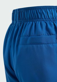 Blue athletic shorts featuring an elastic waistband, smooth fabric, and a single back pocket with stitched trim.